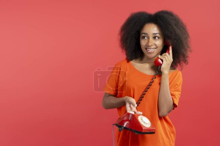 Photo for Portrait of smiling beautiful African American woman with curly hair holding landline red phone, looking away isolated on red background. Retro technology concept - Royalty Free Image