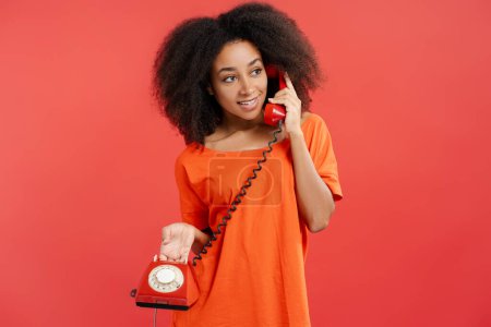 Foto de Retrato de una atractiva mujer afroamericana sonriente con el pelo rizado sosteniendo teléfono fijo rojo, mirando hacia otro lado aislado sobre fondo rojo. Concepto de comunicación - Imagen libre de derechos