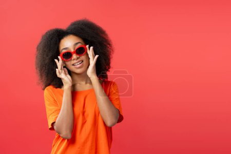 Foto de Retrato de la hermosa mujer joven afroamericana sonriente con el pelo rizado con elegantes gafas de sol rojas aisladas en el fondo rojo, espacio de copia. Concepto de publicidad - Imagen libre de derechos