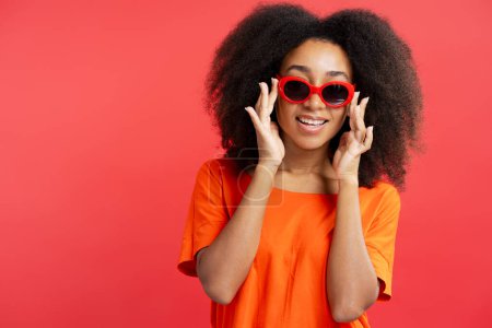 Foto de Mujer joven afroamericana atractiva positiva con el pelo rizado que usa gafas de sol rojas elegantes aisladas en el fondo rojo, espacio de copia, primer plano. Concepto de publicidad - Imagen libre de derechos