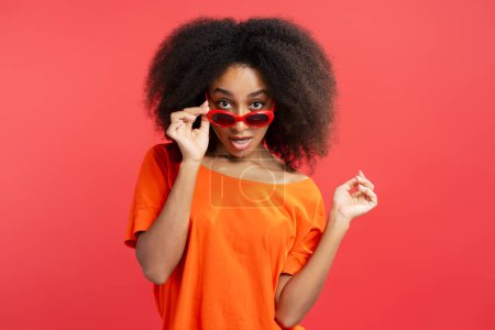 Foto de Modelo femenino afroamericano de moda con cabello rizado con elegantes gafas de sol rojas, mirando a la cámara aislada sobre fondo rojo, espacio para copiar, primer plano. Concepto de publicidad - Imagen libre de derechos