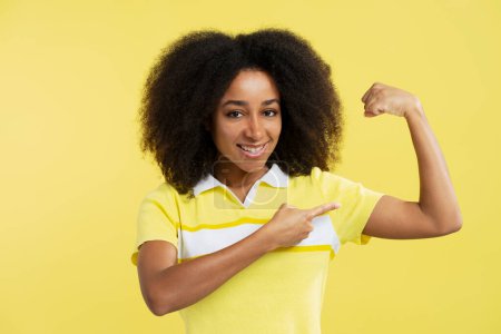 Photo for Portrait of positive African American woman wearing stylish casual clothes pointing finger at her biceps, muscles looking at camera isolated on yellow background, closeup. Concept of healthy lifestyle - Royalty Free Image