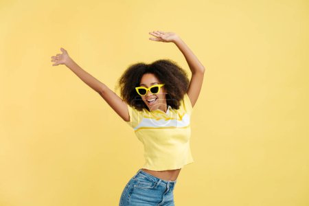 Téléchargez les photos : Portrait de belle jeune femme afro-américaine positive aux cheveux bouclés portant des lunettes de soleil jaunes élégantes isolées sur fond jaune, gros plan. Concept d'été - en image libre de droit