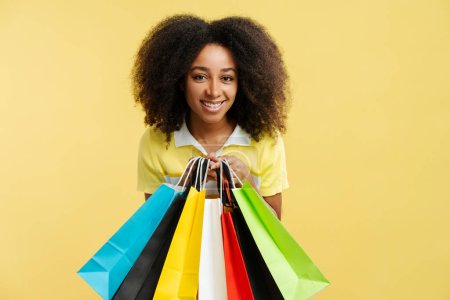 Foto de Retrato de una hermosa mujer afroamericana sonriente sosteniendo coloridas bolsas de compras mirando a la cámara aislada sobre un fondo amarillo. Concepto de compras - Imagen libre de derechos