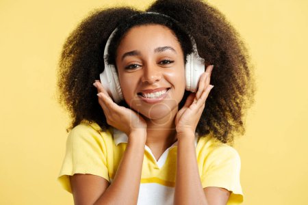 Téléchargez les photos : Portrait d'une belle femme afro-américaine souriante écoutant de la musique dans un casque regardant une caméra isolée sur fond jaune, gros plan. Concept technologique - en image libre de droit