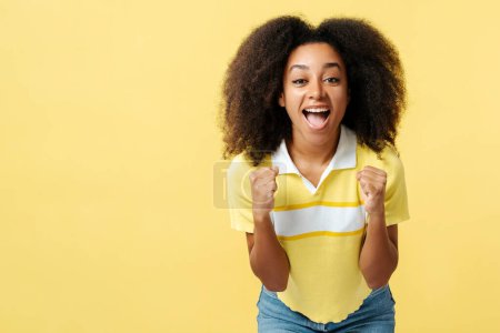 Téléchargez les photos : Portrait de femme afro-américaine excitée aux cheveux bouclés portant des vêtements décontractés gesticulant, tenant les poings, gagnant regardant la caméra isolée sur l'espace de copie de fond jaune. Concept de publicité - en image libre de droit