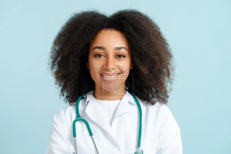 Téléchargez les photos : Portrait de médecin afro-américaine souriante professionnelle en manteau blanc avec stéthoscope regardant la caméra isolée sur fond bleu gros plan. Concept de soins de santé - en image libre de droit