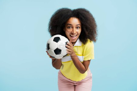 Foto de Retrato de una atractiva mujer afroamericana sonriente con un atuendo casual sosteniendo una pelota de fútbol mirando a la cámara, aislada sobre un fondo azul. Concepto de deporte activo - Imagen libre de derechos