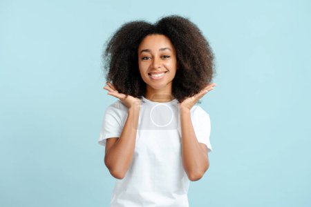 Téléchargez les photos : Heureuse femme afro-américaine authentique aux cheveux bouclés portant un t-shirt blanc regardant la caméra isolée sur fond bleu. Concept de soins capillaires, traitement - en image libre de droit