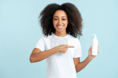 Foto de Retrato sonriente mujer afroamericana con el pelo rizado en camiseta blanca sosteniendo botella mockup dedo apuntando mirando a la cámara aislada en mockup fondo azul. Concepto de publicidad, cuidado del cabello - Imagen libre de derechos