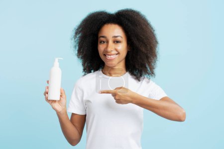 Photo for Portrait happy african american woman with curly hair in white t shirt holding bottle mockup pointing finger looking at camera isolated on blue background. Concept of advertising, cosmetic procedures - Royalty Free Image