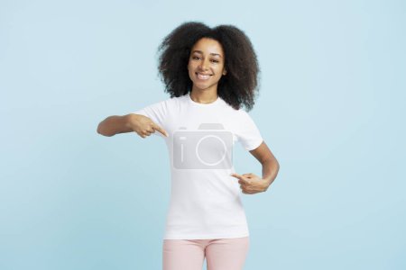 Foto de Retrato de una auténtica joven afroamericana con el pelo rizado vistiendo un atuendo casual, señalando con el dedo la camiseta blanca, maqueta, mirando a la cámara aislada sobre fondo azul. Concepto de publicidad - Imagen libre de derechos