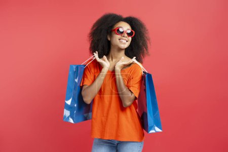 Photo for Young smiling curly haired African American woman wearing stylish eyeglasses holding blue shopping bags looking away isolated on red background. Shopping, black Friday concept - Royalty Free Image