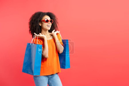 Téléchargez les photos : Jeune femme afro-américaine aux cheveux bouclés heureux portant des lunettes élégantes tenant des sacs à provisions bleus regardant loin isolé sur fond rouge, maquette. Shopping, centre commercial, concept Black Friday - en image libre de droit