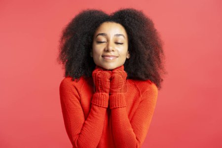 Téléchargez les photos : Belle femme afro-américaine souriante avec les yeux fermés portant des gants d'hiver et un pull chaud isolé sur fond rouge - en image libre de droit