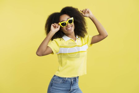 Téléchargez les photos : Portrait de positive attrayante jeune femme afro-américaine aux cheveux bouclés portant des lunettes de soleil jaune élégant isolé sur fond jaune, gros plan. Concept de publicité - en image libre de droit