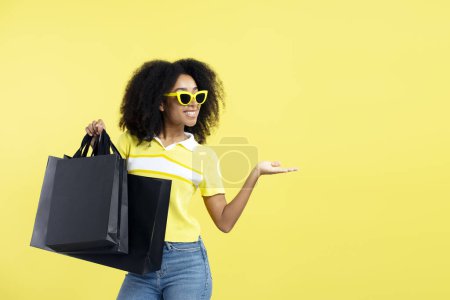 Téléchargez les photos : Belle femme afro-américaine aux cheveux bouclés souriants portant des lunettes de soleil élégantes tenant des sacs à provisions noirs isolés sur fond jaune. Magasin, shopping, vente, concept Black Friday - en image libre de droit