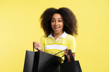 Téléchargez les photos : Jeune femme afro-américaine excitée tenant des sacs à provisions noirs regardant la caméra isolée sur fond jaune. Shopping, vente, concept Black Friday - en image libre de droit