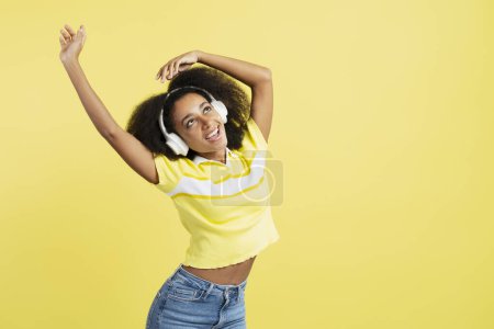 Téléchargez les photos : Heureuse femme afro-américaine portant des vêtements décontractés écoutant de la musique dans des écouteurs dansant en regardant loin isolé sur fond jaune, s'amusant. Concept de publicité - en image libre de droit