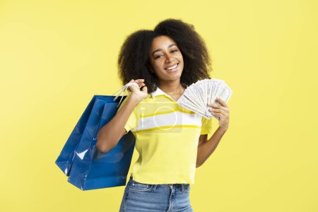 Téléchargez les photos : Belle femme afro-américaine souriante tenant de l'argent et sac à provisions isolé sur fond jaune. Magasin, shopping, vente, concept Black Friday - en image libre de droit