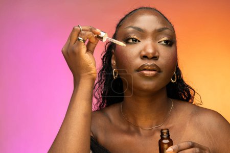 Téléchargez les photos : Portrait rapproché de jeune femme afro-américaine appliquant du sérum à l'huile de pipette, isolé sur fond coloré. Concept de soins du visage - en image libre de droit