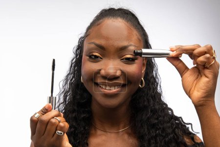 Photo for Smiling African American woman holding mascara, doing makeup, isolated on white background. Beauty and cosmetics concept - Royalty Free Image