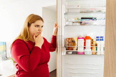 Téléchargez les photos : Belle femme positive corps ouvert réfrigérateur, en choisissant la nourriture dans la cuisine dans un appartement confortable. Concept de perte de poids, mode de vie sain - en image libre de droit