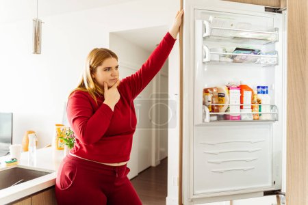 Téléchargez les photos : Pensif, attrayant Caucasien plus la taille femme a ouvert le réfrigérateur, en choisissant la nourriture dans la cuisine dans un appartement confortable. Concept de perte de poids, une alimentation saine - en image libre de droit