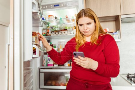 Photo for Portrait of pensive upset young woman standing near open refrigerator, holding mobile phone, making online order at home, in living room. Delivery concept, healthy lifestyle - Royalty Free Image