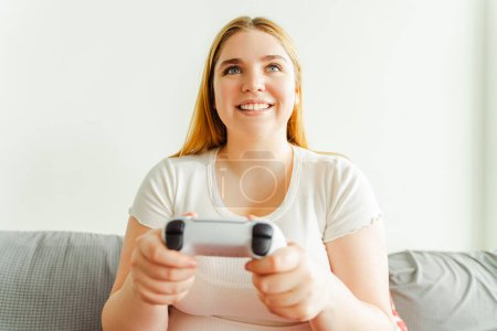 Téléchargez les photos : Portrait de femme séduisante jouant au jeu vidéo, tenant un joystick, assise sur un canapé confortable dans le salon à la maison, gros plan. Concept technologique - en image libre de droit