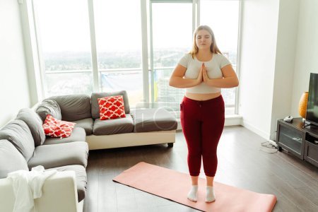 Foto de Atractiva joven mujer de pie con los ojos cerrados en el yoga estera en casa en el apartamento. Concepto de meditación, armonía, equilibrio - Imagen libre de derechos
