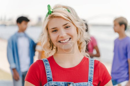 Foto de Retrato de una linda adolescente sonriente con camiseta roja mirando a la cámara en la calle con amigos en el fondo. Estilo de vida positivo, concepto de verano - Imagen libre de derechos