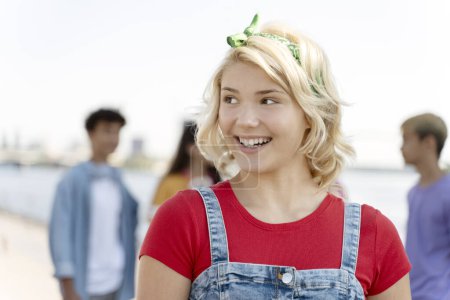 Foto de Retrato de una adolescente sonriente con una camiseta roja mirando hacia la calle con amigos de fondo - Imagen libre de derechos