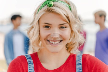 Foto de Retrato de cerca de una adolescente sonriente con una camiseta roja mirando a la cámara en la calle con amigos de fondo. Estilo de vida positivo, concepto de verano - Imagen libre de derechos
