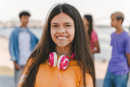 Foto de Retrato de una hermosa adolescente sonriente en camiseta naranja con auriculares mirando a la cámara en la calle, primer plano. Niño auténtico alegre, vacaciones. Concepto de amistad, estilo de vida positivo - Imagen libre de derechos