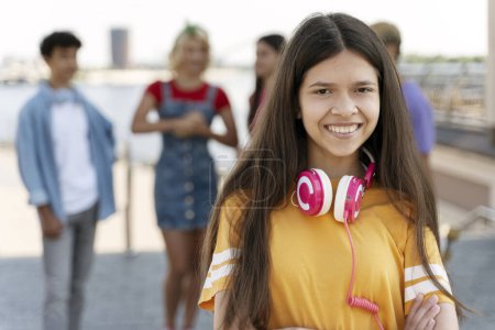 Foto de Retrato de una linda adolescente sonriente con auriculares, camiseta amarilla mirando a la cámara en la calle con amigos en el fondo - Imagen libre de derechos