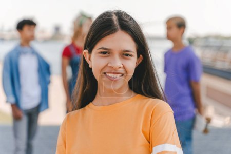 Foto de Retrato de una adolescente sonriente con una elegante camiseta amarilla mirando a la cámara de pie en la calle con amigos de fondo. Verano, concepto de estilo de vida positivo - Imagen libre de derechos