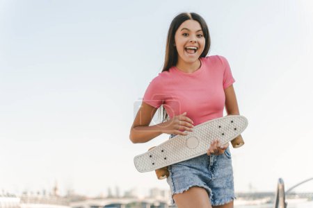 Photo for Portrait of cute emotional teenage girl holding skateboard having fun looking at camera standing on the street. Positive lifestyle, summer concept - Royalty Free Image