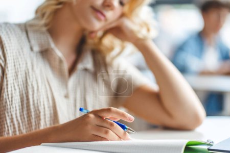 Photo for Pensive teenage girl holding pen, taking notes, sitting at desk in classroom. Attractive schoolgirl doing homework, preparing for exam. Back to school concept - Royalty Free Image