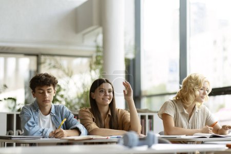 Photo for Smart smiling teenage student raising hand answering question sitting in classroom. Group of teenagers studying together, learning language, exam preparation in modern library. Education concept - Royalty Free Image