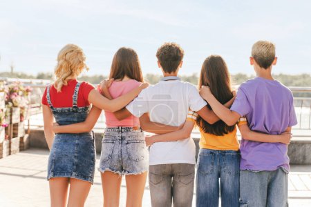 Photo for Back view group of friends, multiracial teenagers wearing colorful t shirts hugging each other looking away standing on the street. Friendship concept - Royalty Free Image