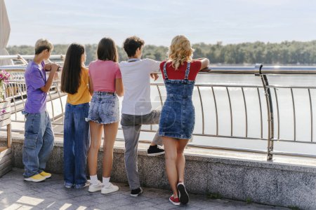 Photo for Back view, positive teenage boys and girls standing by urban street railing outdoors. Friends meeting for walking, communication. Concept of relaxation, summer vacation - Royalty Free Image