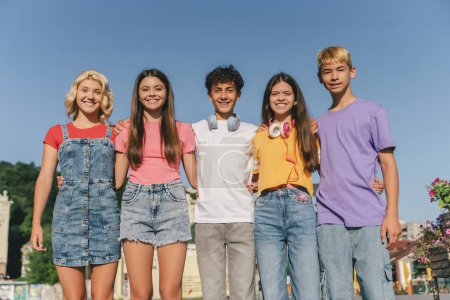 Téléchargez les photos : Groupe d'amis souriants, adolescents multiraciaux portant des t-shirts colorés étreignant debout à l'extérieur. Joyeux garçons et filles élégants dans la rue regardant la caméra. Amitié, mode de vie positif, été - en image libre de droit