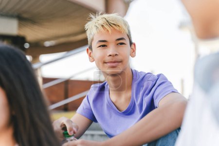 Téléchargez les photos : Portrait d'adolescent souriant élégant avec des cheveux colorés tenant bouteille avec boisson regardant loin assis avec des amis dans la rue urbaine. Jeunesse, concept d'été - en image libre de droit
