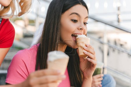 Foto de Retrato de una adolescente sonriente comiendo sabroso helado sentado en la calle con amigos. Concepto de verano - Imagen libre de derechos