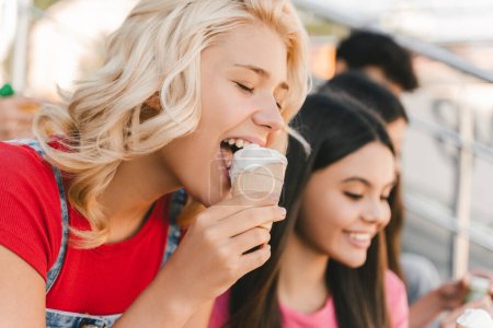 Téléchargez les photos : Adolescente blonde souriante mangeant une délicieuse crème glacée assise dans la rue avec des amis. Concept d'été - en image libre de droit