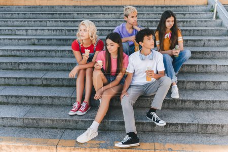 Foto de Grupo de amigos tristes e infelices, adolescentes multirraciales comiendo helado, bebiendo limonada sentados en las escaleras - Imagen libre de derechos