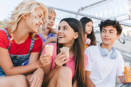 Photo for Group of smiling friends, multiracial teenagers eating ice cream, drinking lemonade sitting outdoors. Happy stylish boys and girls communication on the street. Friendship, positive lifestyle, summer - Royalty Free Image