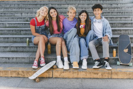 Grupo de amigos sonrientes, elegantes adolescentes multirraciales sentados en las escaleras con patinetas. Feliz elegante comunicación de niños y niñas, relajándose en la calle. Amistad, estilo de vida positivo, verano 