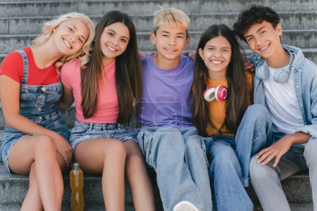 Photo for Group of smiling friends, stylish multiracial teenagers sitting on stairs looking at camera . Happy boys and girls communication, relaxing on the street. Friendship, positive lifestyle, summer - Royalty Free Image